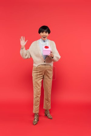 full length of excited asian woman in stylish clothes holding present and waving hand on coral background