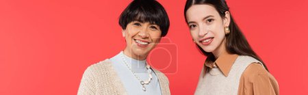 portrait of brunette asian mother and daughter in stylish clothes smiling at camera isolated on coral, banner