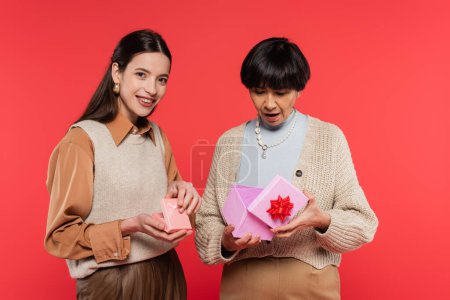 Foto de Surprised asian woman opening gift box near young daughter smiling at camera isolated on coral - Imagen libre de derechos