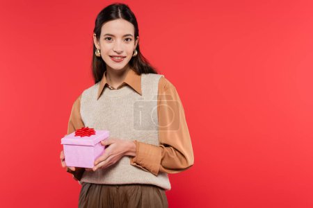 Photo for Happy brunette woman in golden earrings and stylish clothes holding gift box with red bow isolated on coral - Royalty Free Image