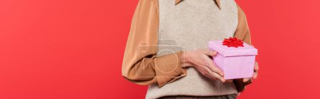 Foto de Cropped view of woman in shirt and knitted vest holding gift box with red bow isolated on coral, banner - Imagen libre de derechos