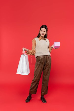 volle Länge der glücklichen Frau in trendiger Freizeitkleidung posiert mit Einkaufstaschen und Geschenkschachtel auf Korallenhintergrund