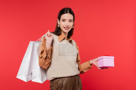 Foto de Young woman in stylish casual attire holding present and shopping bags while smiling at camera isolated on coral - Imagen libre de derechos