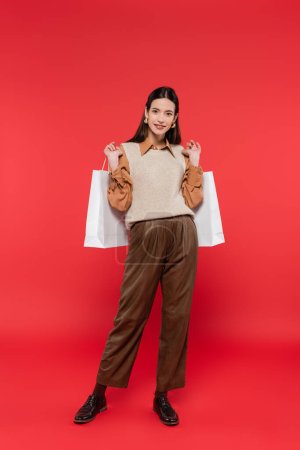 Téléchargez les photos : Full length of smiling woman in brown pants and knitted vest standing with white shopping bags on coral background - en image libre de droit