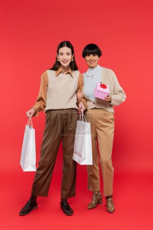 volle Länge der freudigen asiatischen Mutter und Tochter in stilvollen Kleidern stehend mit Geschenkbox und Einkaufstaschen auf Korallenhintergrund