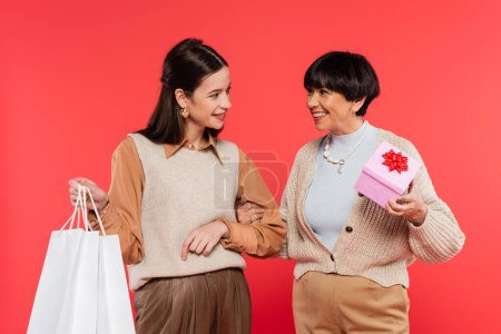happy asian mother and daughter with gift box and shopping bags looking at each other isolated on coral