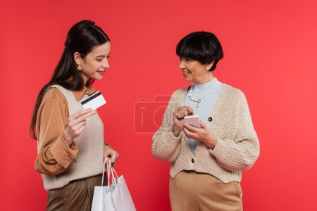 smiling asian woman pointing at smartphone near young daughter holding shopping bags and credit card isolated on coral