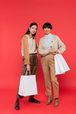 full length of trendy asian mother and daughter posing with white shopping bags on coral background