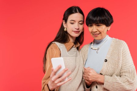 cheerful asian mother and daughter looking at smartphone isolated on coral 
