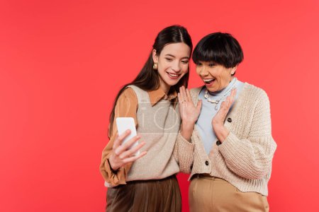cheerful and young asian daughter holding smartphone near excited mother isolated on coral 