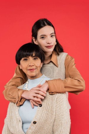 young asian daughter in trendy clothes hugging mother isolated on coral 