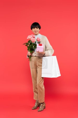 full length of mature asian woman holding flowers and shopping bags on coral background 