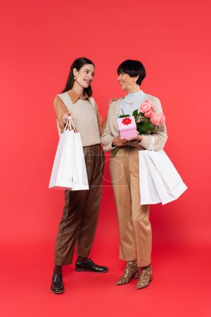 full length of young asian daughter with shopping bags looking at happy mother holding flowers and gift box on coral