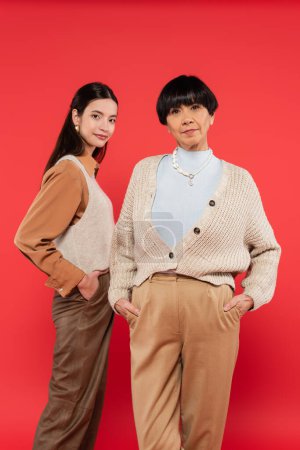 stylish asian mother and daughter in trendy clothes posing with hands in pockets on coral background 