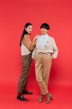 full length of stylish asian woman in trendy clothes standing near mother posing with hands in pockets on coral background 