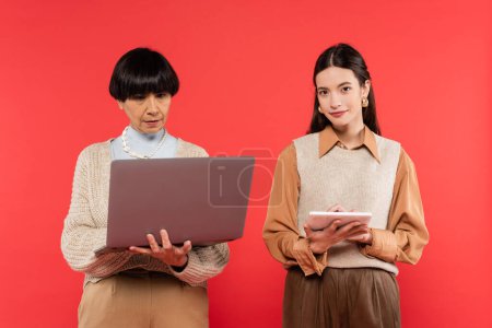 middle aged woman using laptop near young asian daughter holding digital tablet isolated on coral 