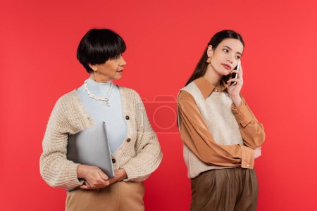 young asian woman talking on smartphone while mother holding laptop isolated on coral 