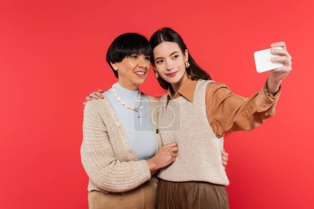 young asian woman in knitted vest taking selfie with happy mother isolated on coral 