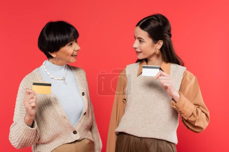 cheerful asian mother and daughter in stylish clothes holding credit cards isolated on coral 