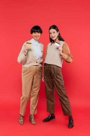 full length of happy asian mother and daughter in stylish clothes holding credit cards on coral background 