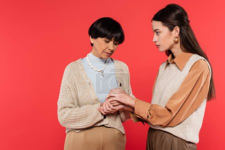 worried asian woman holding hands of upset middle aged mother isolated on coral 