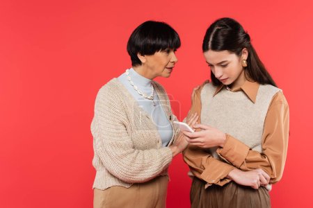 middle aged mother talking to concentrated asian daughter using smartphone isolated on coral 