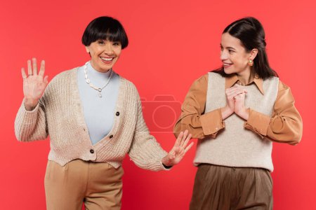 cheerful asian daughter in stylish clothes looking at excited mother waving hands on coral background 