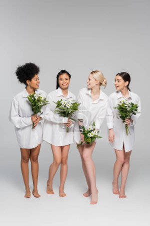 full length of barefoot multicultural women in white shirts holding bouquets on grey background