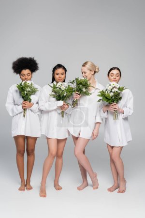 Photo for Full length of young women with bare legs wearing white shirts and enjoying flavor of aromatic flowers on grey background - Royalty Free Image