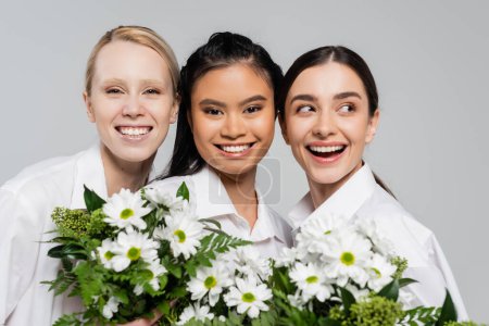 jóvenes mujeres multiétnicas en camisas blancas sonriendo cerca de flores blancas con hojas verdes aisladas en gris