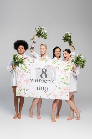 Foto de Longitud completa de alegres mujeres multiétnicas con flores y pancarta de saludo con letras de día para mujer sobre fondo gris - Imagen libre de derechos