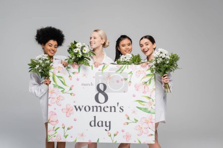 junge interrassische Frauen lächeln in der Nähe von Blumen und riesigen Grußkarten zum Frauentag isoliert auf grau