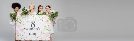 young multicultural women smiling near flowers and mothers day greeting placard isolated on grey, banner