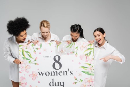 Erstaunte Frau zeigt auf riesige Grußkarte zum Frauentag in der Nähe junger multiethnischer Models, isoliert auf grau