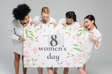 glückliche multiethnische Frauen in weißen Hemden, die eine riesige Grußkarte zum Frauentag betrachten, isoliert auf grau