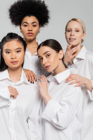 Foto de Pretty multiethnic women wearing white shirts and looking at camera isolated on grey - Imagen libre de derechos