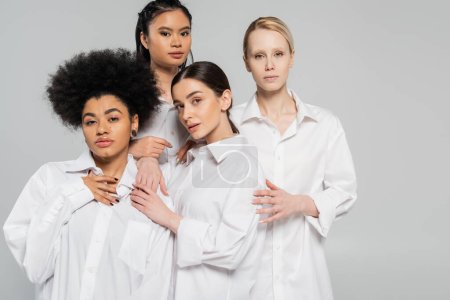 Foto de Sensual multicultural women in white shirts looking at camera while standing isolated on grey - Imagen libre de derechos