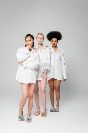 full length of multiethnic women in white shirts and slippers standing on grey background
