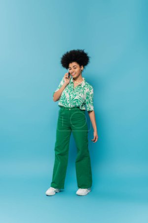 full length of happy african american woman in green pants and leaf print blouse talking on mobile phone on blue background