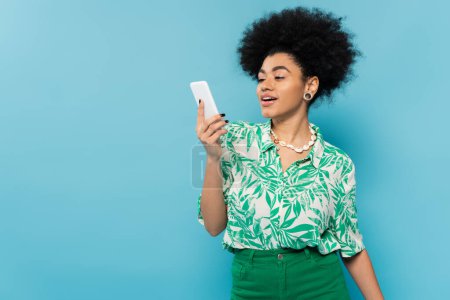 mujer afroamericana positiva en blusa estampada de hoja mirando el teléfono móvil aislado en azul