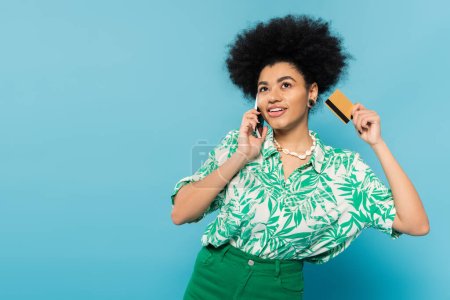 african american woman in stylish blouse holding credit card and talking on smartphone isolated on blue