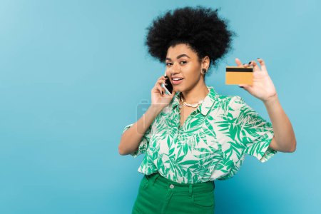 mujer afroamericana de moda mostrando crédito y hablando por teléfono celular mientras sonríe a la cámara aislada en azul