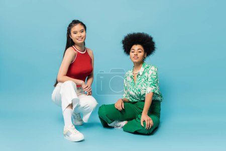 full length of trendy multiethnic women in trendy outfit posing and smiling at camera on blue background