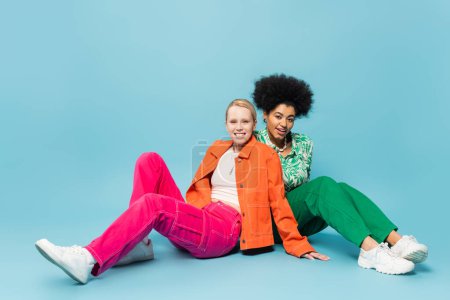 full length of blonde and brunette interracial women in bright and trendy attire sitting on blue background