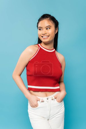 joyful asian woman in red top looking away while holding hands in pockets of white pants isolated on blue