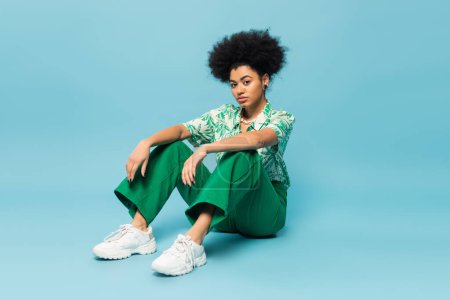 full length of curly african american woman in green pants and white sneakers sitting on blue background