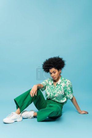 full length of african american woman in stylish blouse and green pants sitting and looking at camera on blue background