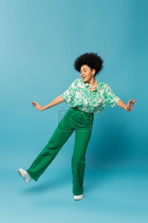 full length of cheerful african american woman in leaf print blouse and green pants posing on blue background