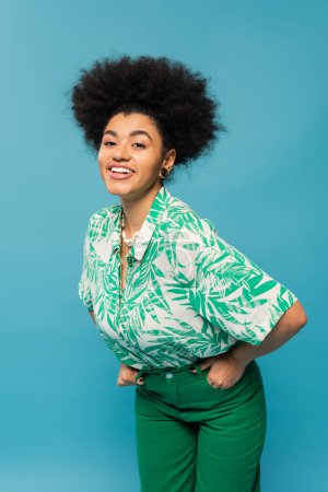 trendy african american woman in leaf print blouse smiling at camera isolated on blue