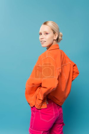 young blonde woman in bright orange jacket posing with hand on hip and looking at camera isolated on blue